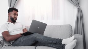 Online communication. Flirting man. Home relax. Handsome arabian guy sitting comfortable sofa typing laptop in light room interior.