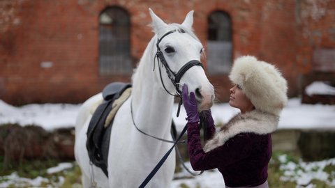 horsewoman is communication with white horse in horse yard in winter, beautiful woman