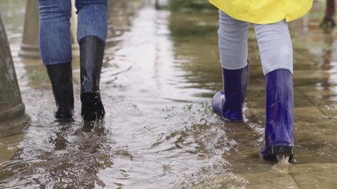 people walk along road in rubber boots through puddles, splashes fly from walking, wet asphalt road, flood city street, water spilling from banks, earth weather phenomenon, mud and puddles underfoot