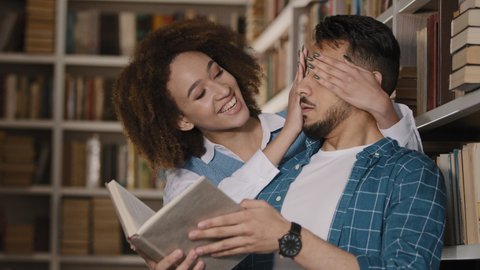 Young hispanic male student stands in library reading book african american girl unexpectedly approaches guy closes eyes surprises friends hugging smiling talking communicate happy to see each other
