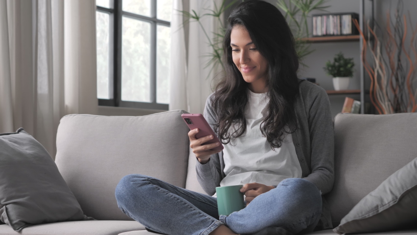 Cheerful young arab woman sitting cross legged on the couch using smart phone mobile on internet,latin girl sits on the sofa holding smartphone searching online | Shutterstock HD Video #1085420324