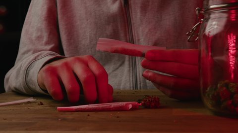 Caucasian male hands making a cigarette with marijuana. Red and blue light illuminated hands and glass jar with CBD buds on the table.