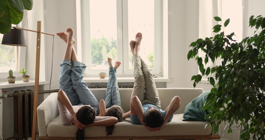 Rear view young couple and little 5s preschool daughter lying down on cozy sofa in living room having fun raise move their barefoot feet enjoy active playtime at home. Family weekend, leisure concept Royalty-Free Stock Footage #1085422394