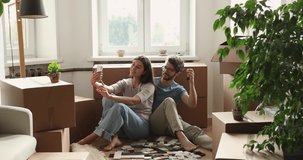 Happy couple sit on floor near cardboard boxes with personal stuff, use smart phone take selfie, show keys, make picture to remember day of moving to own house. Tenancy, modern tech, relocate concept