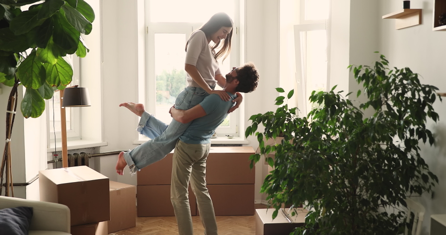 Husband lifts on hands beloved wife, happy millennial couple homeowner family celebrate relocation day to own or rented house, houseplant, cardboard boxes nearby. Bank loan, mortgage, move day concept Royalty-Free Stock Footage #1085422883