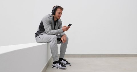 Man listening to podcast, audiobook or music on mobile phone app wearing headphones sitting at home. Healthy lifestyle sport athlete using smartphone on running break outdoors. Man in sweatpants