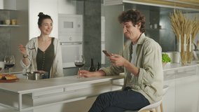 Charming woman cooking some tasty meal on kitchen and giving her husband to taste it on spoon. Handsome man sitting near with smartphone in hands and drinking wine. - Powered by Shutterstock - Get 15% off with code: PIKWIZARD15