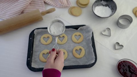 Woman sieving icing sugar over heart shaped biscuits on a baking tray. Baking food treats concept with rolling pin, cookie cutters, jam and biscuits in frame. White background. Real time 4k motion 