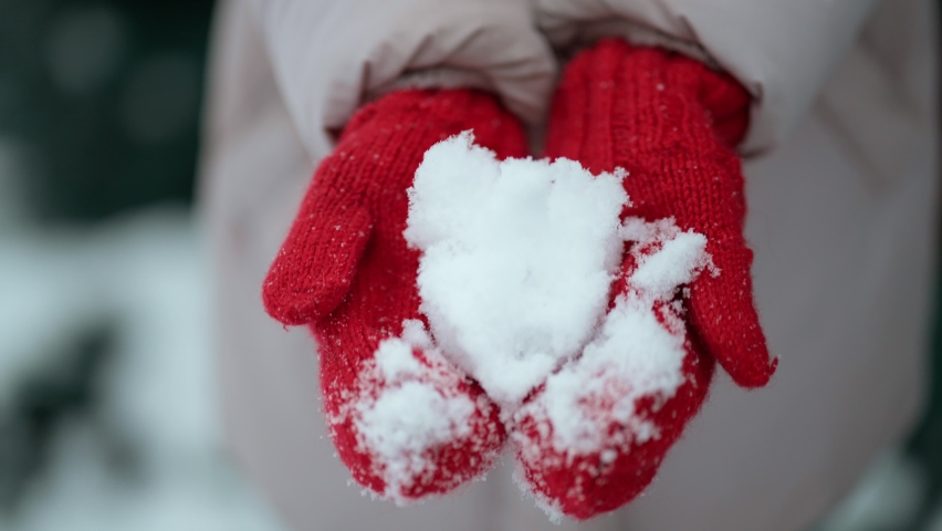 Snow like heart shape in hands. Charming woman holds snow in hands in warm red mittens, puts hands up to face and looks at camera through snow. Valentine