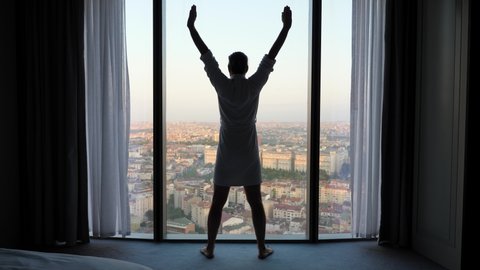 Man finishes his morning exercise, raises his arms to his sides and up and then down. Silhouette of a man standing in front of a window, city panorama visible outside, apartments bedroom at top floor