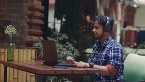 Moving shot of a modern young Indian Asian male wearing headphones working remotely through a video conference call using a laptop and headset sitting in an outdoor sidewalk cafe and restaurant.