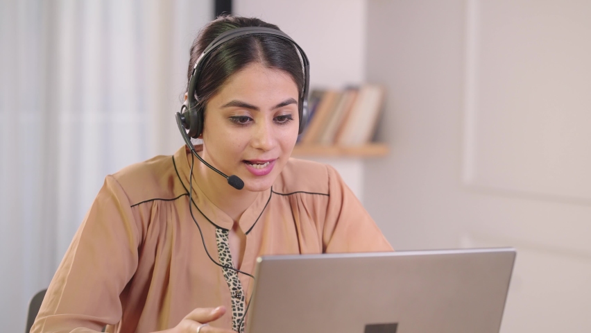 Smiling young attractive Indian Asian female corporate call center employee wearing headphones having a conversation with a client or customer working at a desk at workspace or office using a laptop.  Royalty-Free Stock Footage #1085437709