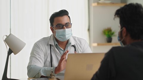 Indian Asian male doctor sitting in a modern clinic wearing stethoscope and apron giving consultation to a female patient, both wearing protective mask on face. Medical, medicine, healthcare concept