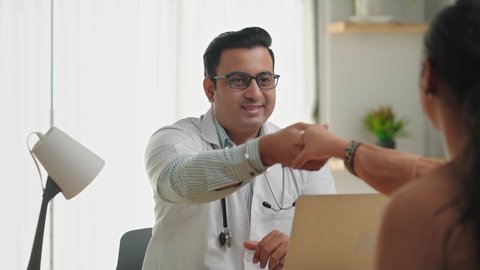 An Indian Asian male smiling physician or doctor sitting in a modern clinic wearing a stethoscope and apron shaking hand with a female patient customer. Medical, medicine, and healthcare concept