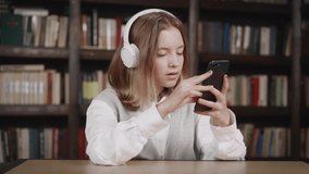 Cute caucasian kid girl holding smartphone enjoying using mobile apps, playing games sitting in library. Child learning in cell phone, watching video, having fun with mobile technology concept
