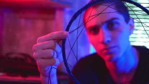 Artisan makes new dreamcatcher in art studio. Master in dark workshop with neon red and blue lighting, close up, soft selective focus.