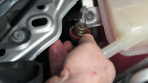 Close-up footage of a mechanic's hand slipping into the engine compartment of a car and removing a headlight bulb to replace. Concept of repair and maintenance, do it yourself.