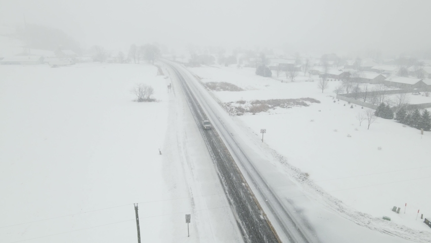 Snow storm in Wisconsin valley. Single vehicle on the highway. Poor visibility in the distance. Residential neighborhood covered in snow. 