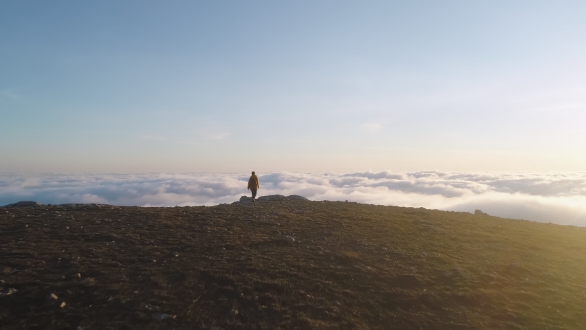 Solo girl with backpack walks on mountain top above clouds and looks at sunset