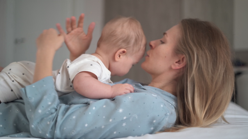beautiful young caucasian woman lies on her back and holds her baby on her stomach. Mom hugs and kisses her little son. Mother