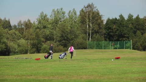 A man and a woman in a pink vest are walking on a golf course and carrying carts with clubs on a sunny day. Summer golf in cool weather.