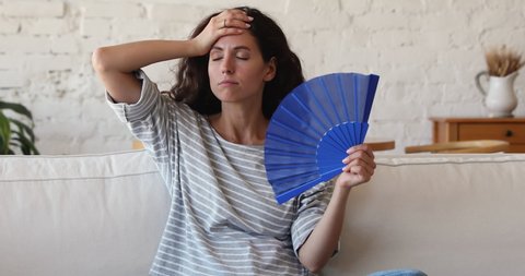 Exhausted 35s Hispanic woman cools herself with blue paper waver fan, sit on sofa in living room, suffers from too hot summer weather, high temperature inside. Discomfort, need air conditioner concept