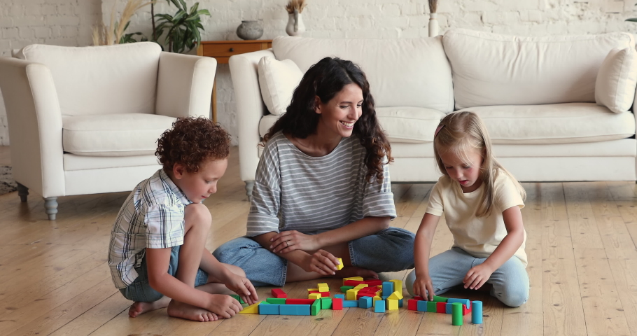 Hispanic babysitter play wooden cubes with diverse boy and girl at home, mother her little son and daughter sit on warm floor in modern cozy living room enjoy playtime. Childhood activity, fun concept Royalty-Free Stock Footage #1085456354