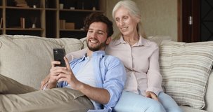 Grownup son spend time with older mum relax on sofa with cellphone in living room. Multi generation family enjoy modern device, watch online videos on cell at home. Leisure, fun, wireless tech concept