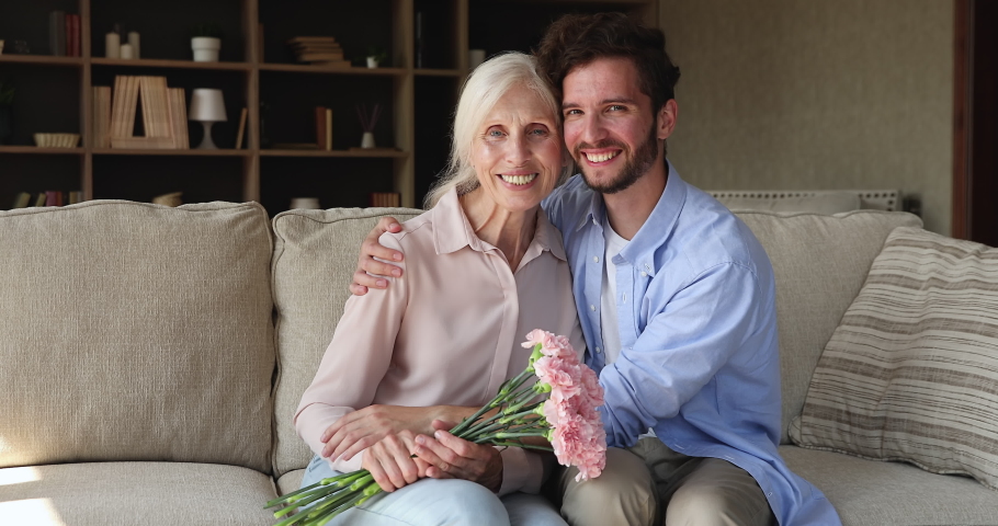 Older woman holds carnations flowers sit on sofa with young adult grown up son smile look at camera. Multi generation family life events celebrations, Happy Mothers Day congrats, bond and care concept Royalty-Free Stock Footage #1085456567
