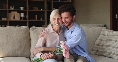 Older woman holds carnations flowers sit on sofa with young adult grown up son smile look at camera. Multi generation family life events celebrations, Happy Mothers Day congrats, bond and care concept