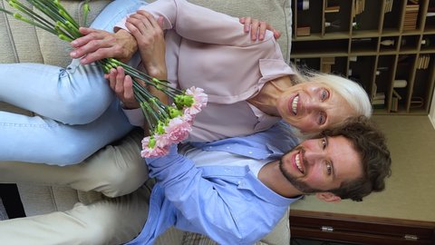 Vertical view happy grown up son cuddles his older mom holds bouquet of carnation. Family celebrate life event, international Mothers or Womens day, anniversary or birthday at home. Love, care concept