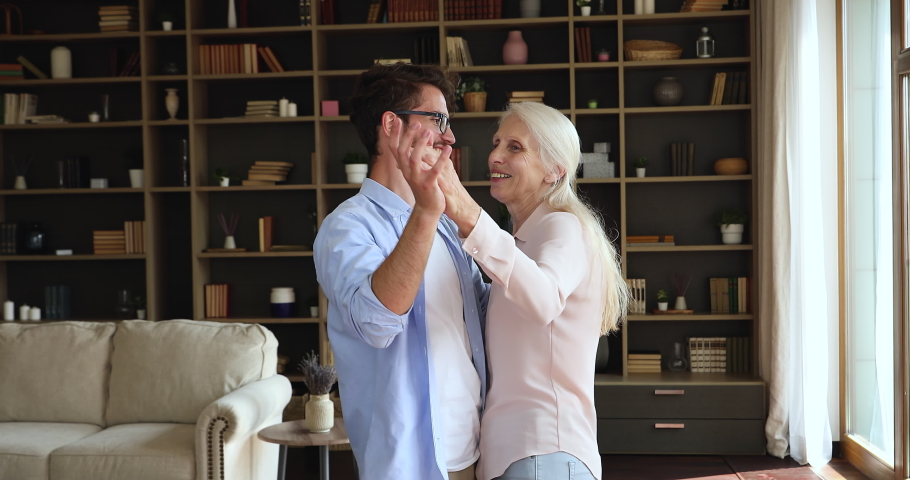 Grey-haired older woman holding hands of grown up young adult son dance waltz enjoy music and carefree weekend leisure together in modern home. Multi generational family bonding, hobby, love concept Royalty-Free Stock Footage #1085456699