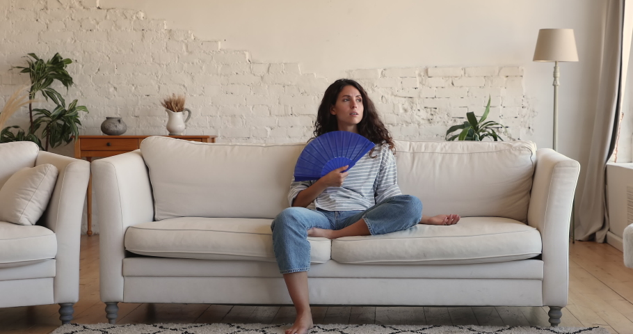 Overheated Latina female sit on sofa in living room at hot summer day feel discomfort suffers from too hot air inside, wave blue fan to cool herself, sweating due house without air conditioner concept Royalty-Free Stock Footage #1085456717