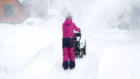 Child in crimson winter overalls and hat among snowdrifts during snowfall in yard of house removes snow with snowplow, back view. Lot of snow. Slow motion, 4k.