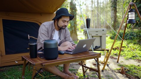Concentrated Asian man using laptop working remotely while camping in the forest. work and vacation concept.