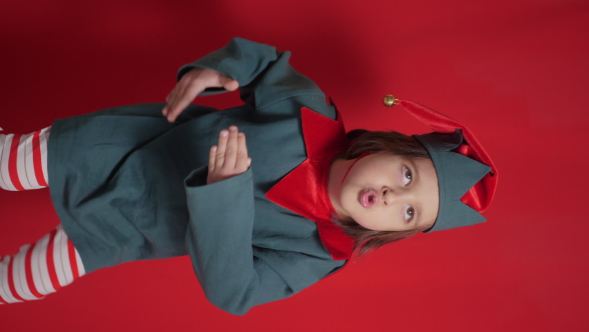 baby boy in a green suit and a dwarf hat is indulging on a red background for Christmas vertical