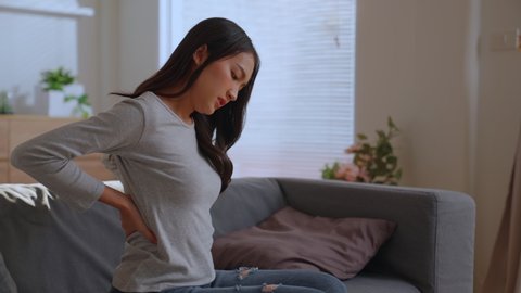 Charming asian woman sitting on sofa pressing hands against lower back, feels pain suffers from injury back arthritis membrane holding on to spine. back pain, office syndrome, stretching morning.