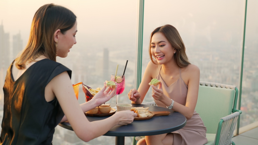 4K Young Asian woman friends celebrating dinner party meeting at skyscraper rooftop restaurant bar in metropolis at summer sunset. Beautiful girl enjoy outdoor lifestyle together on holiday vacation.