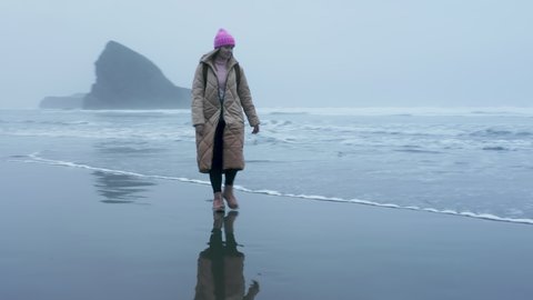 Cinematic woman silhouette reflection on wet beach surface in Oregon coast. Drone flying around woman traveler with backpack walking by ocean beach. Epic aerial flying around person low above water 4K