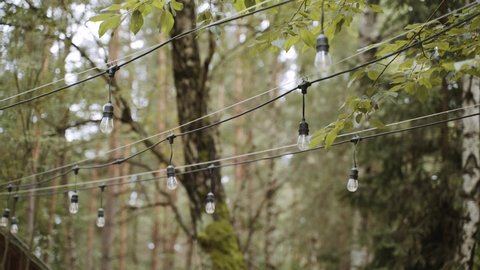 Switched off garland of light bulbs on black wire hangs between green trees on a summer day. Home yard decoration.