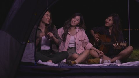 Friendship group of Asian young women having fun and enjoy music form guitar in tent. mountain outdoor camping trip in nature at night. Females Relaxation, Lifestyle, Vacations 