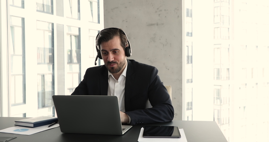 Virtual meeting by business, distance communication, negotiations event concept. Millennial businessman wear headset sit at workplace use laptop talks on videoconference app to client, assist remotely Royalty-Free Stock Footage #1085474840