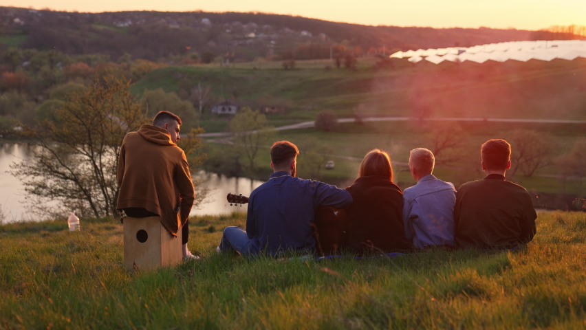 Four young men and woman having good picnic time. Friendly group watching the sunset and singing song along with playing instruments.