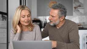 Cheerful middle-aged couple with a laptop sitting at the desk in modern kitchen at home, choosing new furniture, making food order, scrolling news feed. Mature spouses spend leisure time together - Powered by Shutterstock - Get 15% off with code: PIKWIZARD15