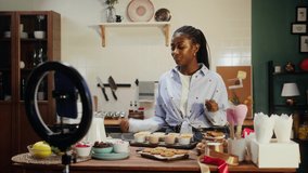 African american chef blogger cooking cupcakes and recording video on smartphone camera. Woman confectioner showing how to prepare muffins, tasty dessert. Baking sweet food, bakery at home. 