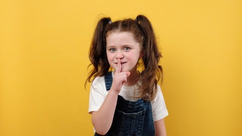Portrait of cute beautiful little child girl saying hush be quiet with finger on lips shhh gesture, posing isolated over yellow color background wall in studio. People childhood lifestyle concept