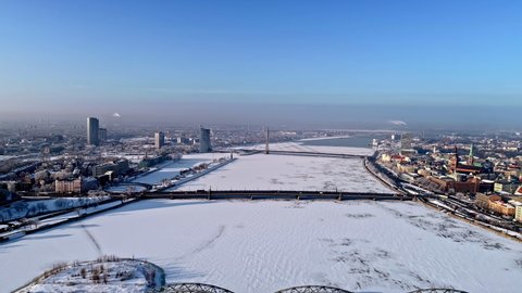 Aerial flyover snow-covered Daugava River during cold winter day with sun and blue sky - Cars crossing bridge to City of Riga,Latvia