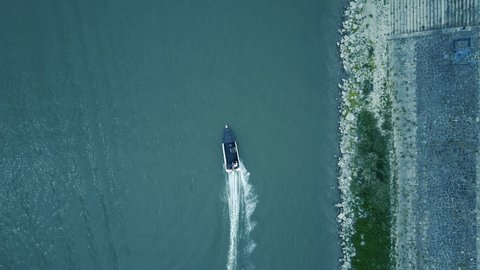 A black and white boat, speedboat racing across the picture, skid slight left turn, just below the drone, aerial shot, blue water and tidy rocky waterfront coast