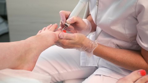 Pedicurist woman removes shellac polish from toes using manicure machine, closeup view. Pedicure Master is removing red gel polish from nails on toes using electric nail drill in cosmetology.