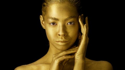 Attractive Female Model With Golden Skin Posing Touching Face And Skin Covered With Glowing Gold Body Paint Or Powder Looking At Camera Over Black Studio Background. Beauty Portrait, Fashion Art